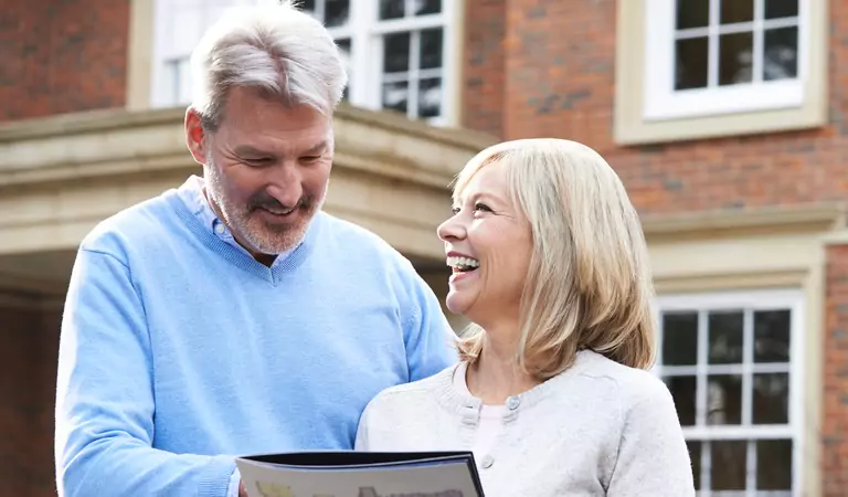 old age couple standing outside of a house and reading something